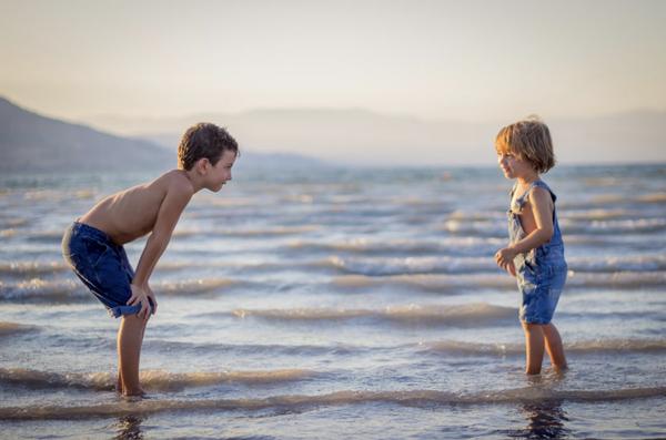boy bending knee while watching younger child on beach